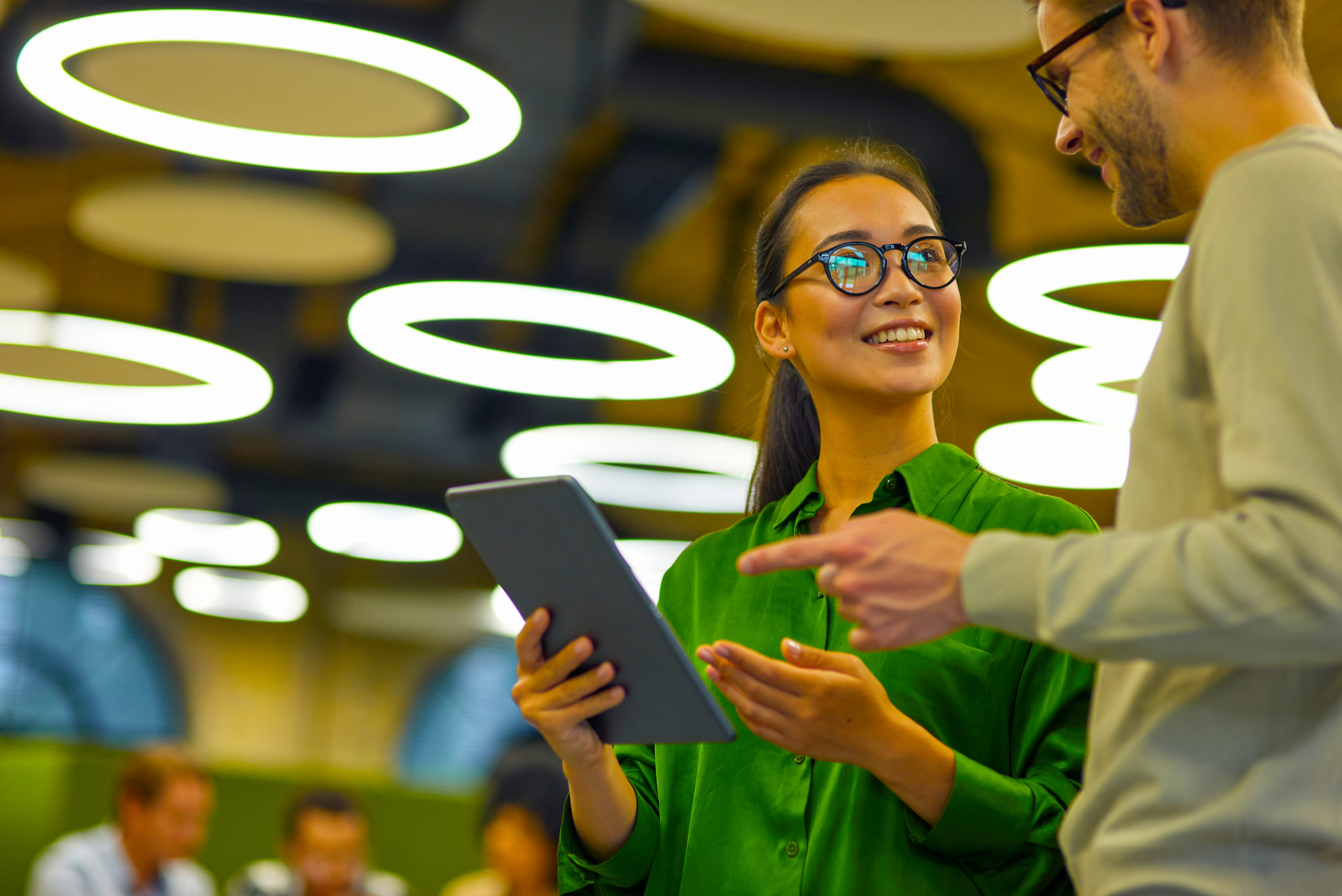 Discussing project. Young positive asian woman showing something on digital tablet to her male colleague while standing together in the coworking space or office