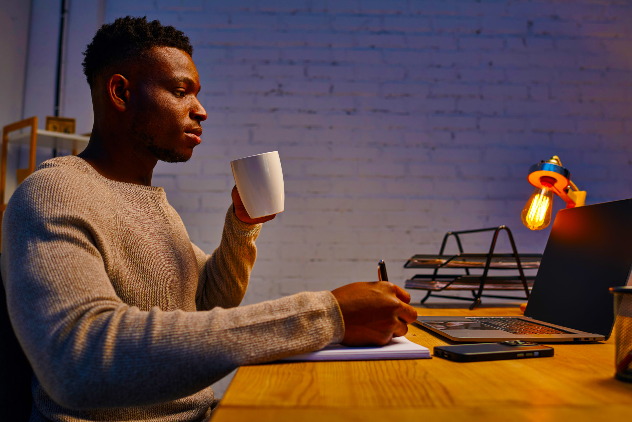 african american guy with coffee cup writing in notebook near laptop working in home office at night